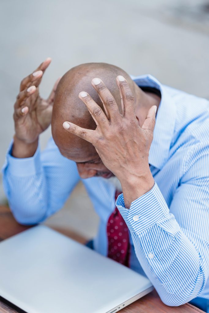 African American male wearing formal outfit grabbing head with hands and looking down at closed laptop while sitting at table