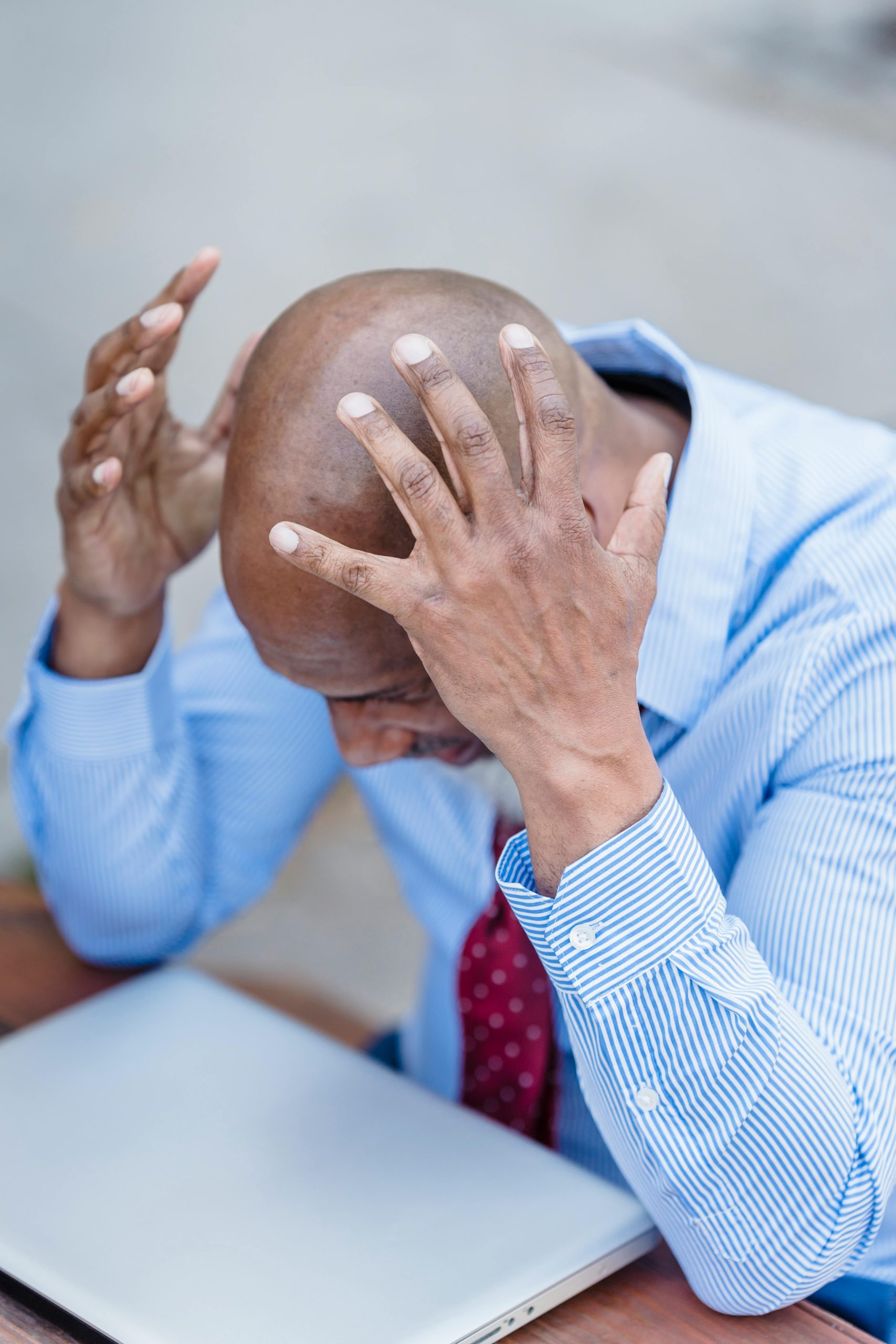African American male wearing formal outfit grabbing head with hands and looking down at closed laptop while sitting at table
